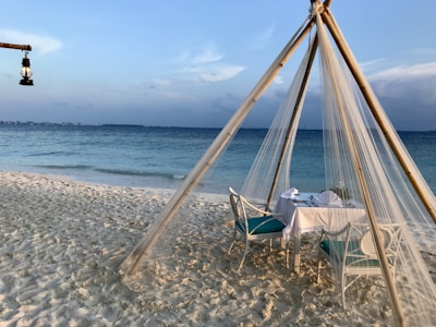 A serene beachfront setting with a dining table under a bamboo and sheer fabric canopy. The sandy beach leads to calm blue ocean waters, with a lantern hanging on the left adding a touch of rustic charm.