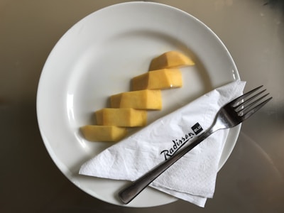 Slices of yellow mango are neatly arranged on a white plate. A silver fork rests on a white napkin, which is placed on the plate, featuring the Radisson Blu logo.