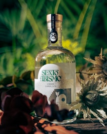 A bottle of gin labeled 'SEXTO ABISMO' is surrounded by lush greenery and flowers. The label features artistic designs, including a cat and abstract graphics.
