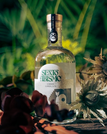 A bottle of gin labeled 'SEXTO ABISMO' is surrounded by lush greenery and flowers. The label features artistic designs, including a cat and abstract graphics.