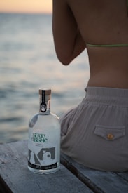 A bottle of liquor sits on a wooden surface near the ocean. The bottle label reads 'Sexto Abismo.' Next to the bottle, a person wearing a swimsuit is seated, facing away from the camera.