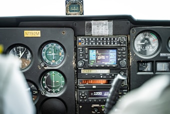 A detailed view of an airplane cockpit instrument panel, featuring various gauges, dials, and controls. Prominently visible are the altimeter, GPS, compass, radio communication devices, and engine RPM gauge. The panel includes a small note taped above one of the instruments.