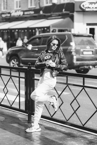 A person in a casual outfit leans against a metal railing on a city street, holding a drink. The individual is wearing sunglasses, a leather jacket, and sneakers, exuding a relaxed and confident demeanor. A parked car and people walking are visible in the background, adding to the urban atmosphere.
