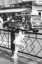 A person in a casual outfit leans against a metal railing on a city street, holding a drink. The individual is wearing sunglasses, a leather jacket, and sneakers, exuding a relaxed and confident demeanor. A parked car and people walking are visible in the background, adding to the urban atmosphere.