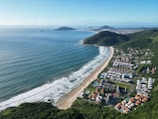 A coastal landscape features a long stretch of beach bordered by the ocean on one side and dense green hills on the other. Residential buildings and some recreational areas with pools are visible near the beach. The horizon shows additional hills and islands in the distance under a clear blue sky.