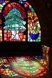 A vibrant stained glass window featuring an abstract design with a large central tree and a flower in a pot. The sunlight filtering through the glass projects a colorful pattern onto the floor, creating a mosaic of colors and shapes on the surface.