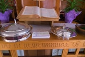 An open book placed on a wooden stand, flanked by two silver-colored, lidded containers with crosses on top. A stack of pamphlets or cards is positioned in front of the containers. Two potted plants with green leaves and purple wrapping are visible in the background. The wood surface has engraved text.