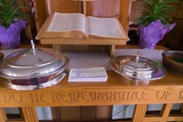 An open book placed on a wooden stand, flanked by two silver-colored, lidded containers with crosses on top. A stack of pamphlets or cards is positioned in front of the containers. Two potted plants with green leaves and purple wrapping are visible in the background. The wood surface has engraved text.