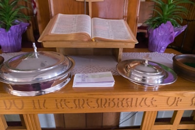 An open book placed on a wooden stand, flanked by two silver-colored, lidded containers with crosses on top. A stack of pamphlets or cards is positioned in front of the containers. Two potted plants with green leaves and purple wrapping are visible in the background. The wood surface has engraved text.