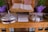 An open book placed on a wooden stand, flanked by two silver-colored, lidded containers with crosses on top. A stack of pamphlets or cards is positioned in front of the containers. Two potted plants with green leaves and purple wrapping are visible in the background. The wood surface has engraved text.
