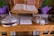 An open book placed on a wooden stand, flanked by two silver-colored, lidded containers with crosses on top. A stack of pamphlets or cards is positioned in front of the containers. Two potted plants with green leaves and purple wrapping are visible in the background. The wood surface has engraved text.