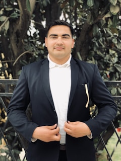 A person stands confidently outdoors wearing a dark suit and white shirt, hands clasped in front of them. The background consists of dense greenery and a fence, suggesting a garden or park setting.