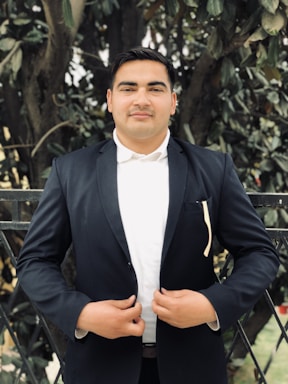 A person stands confidently outdoors wearing a dark suit and white shirt, hands clasped in front of them. The background consists of dense greenery and a fence, suggesting a garden or park setting.