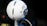 A spherical white microphone is positioned on a stand, featuring silver mesh grills and a prominent logo in the center. The background is softly blurred, emphasizing the microphone's design and the intricate details of its structure.