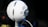 A spherical white microphone is positioned on a stand, featuring silver mesh grills and a prominent logo in the center. The background is softly blurred, emphasizing the microphone's design and the intricate details of its structure.