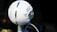 A spherical white microphone is positioned on a stand, featuring silver mesh grills and a prominent logo in the center. The background is softly blurred, emphasizing the microphone's design and the intricate details of its structure.