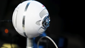 A spherical white microphone is positioned on a stand, featuring silver mesh grills and a prominent logo in the center. The background is softly blurred, emphasizing the microphone's design and the intricate details of its structure.