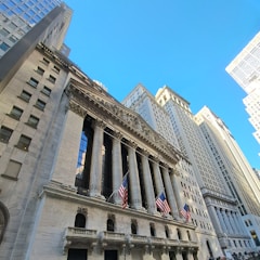 A grand neoclassical building with a colonnaded facade rises against a clear blue sky. Several large American flags are displayed prominently. The building is flanked by tall skyscrapers, creating an impression of being in a financial district.