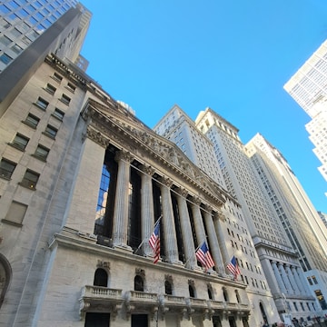 A grand neoclassical building with a colonnaded facade rises against a clear blue sky. Several large American flags are displayed prominently. The building is flanked by tall skyscrapers, creating an impression of being in a financial district.
