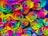 A collection of vibrant multicolored roses with petals in a spectrum of colors including red, orange, yellow, green, blue, and purple. Water droplets are visible on the petals, enhancing the freshness and vividness of the colors.