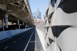 The image features a modern urban landscape with a concrete pathway running between an elevated roadway supported by metal girders and a textured wall with geometric patterns. In the background, a city skyscraper rises against a clear blue sky, suggesting a metropolitan setting.