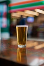 A glass of beer with a golden hue stands on a glossy wooden table, casting a soft reflection. The background features colorful, blurred store shelves and lights, adding to the casual and warm atmosphere.