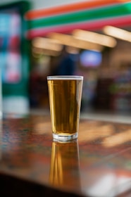 A glass of beer with a golden hue stands on a glossy wooden table, casting a soft reflection. The background features colorful, blurred store shelves and lights, adding to the casual and warm atmosphere.