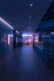A dimly lit hallway leads to an illuminated store entrance with a person standing at the door. The ambiance presents a futuristic, neon-lit environment with soft bluish-purple hues. The store is stocked with various products, visible through the glass windows.