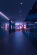 A dimly lit hallway leads to an illuminated store entrance with a person standing at the door. The ambiance presents a futuristic, neon-lit environment with soft bluish-purple hues. The store is stocked with various products, visible through the glass windows.