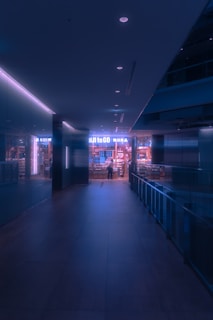 A dimly lit hallway leads to an illuminated store entrance with a person standing at the door. The ambiance presents a futuristic, neon-lit environment with soft bluish-purple hues. The store is stocked with various products, visible through the glass windows.