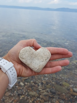A hand holds a heart-shaped stone over a body of water with smooth pebbles visible below the surface. The background features a serene lake extending into the distance under a partly cloudy sky.