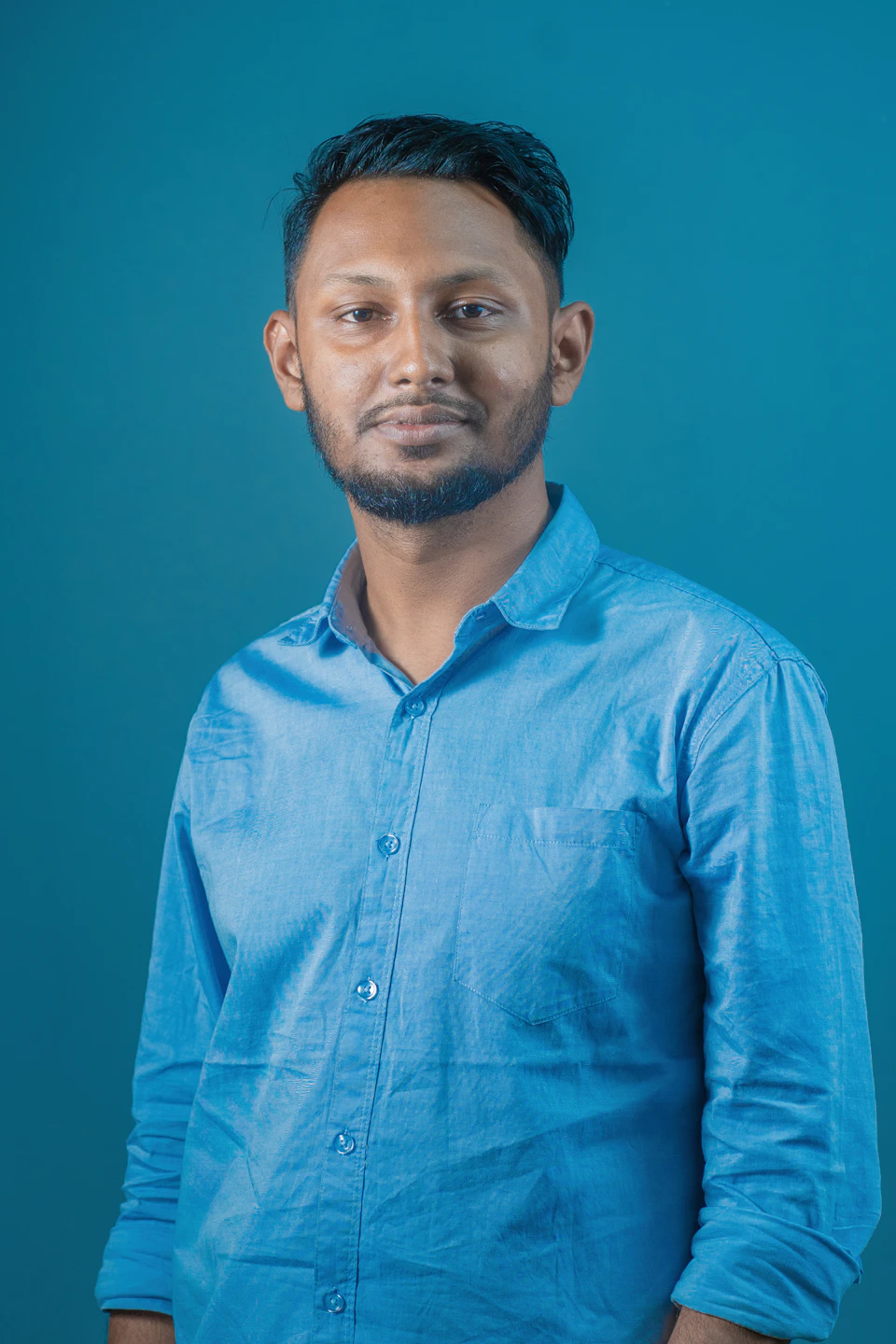 The image shows a man standing against a blue background, wearing a light blue collared shirt and dark hair. The man is looking directly at the camera with an expression of concentration or thoughtfulness on his face.