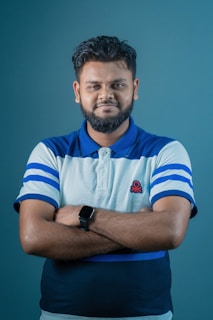 A person is standing with arms crossed, wearing a polo shirt with blue and white stripes and a logo on the chest. They have a short beard and are wearing a smartwatch. The background is a solid teal color, and the lighting is even.