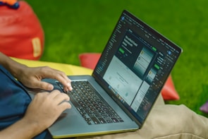 A person is typing on a laptop, which is displaying a website with a dark interface. The setting appears casual with colorful bean bags in the background, resting on a green grass-like surface.