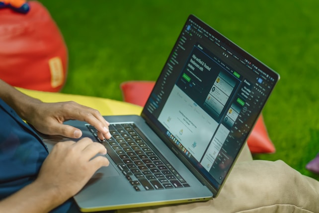 A person is typing on a laptop, which is displaying a website with a dark interface. The setting appears casual with colorful bean bags in the background, resting on a green grass-like surface.