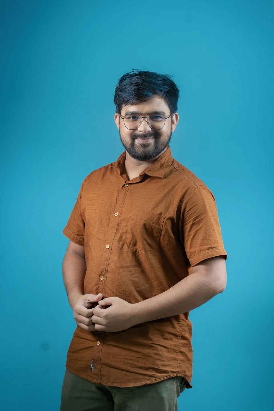 The image shows a man standing against a blue background, wearing an orange shirt and glasses. He is smiling at the camera with his hands in his pockets. The man's attire consists of a short sleeve shirt paired with khaki pants.