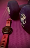 A close-up view of a fitness watch with a red strap placed on a textured purple exercise mat. Beside the watch are two large blue dumbbells with white branding on the ends.