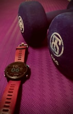 A close-up view of a fitness watch with a red strap placed on a textured purple exercise mat. Beside the watch are two large blue dumbbells with white branding on the ends.