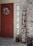 A front porch featuring a red door adorned with a floral wreath. Next to the door is a narrow window with blinds. Leaning against the brick wall is a wooden sign with the word 'WELCOME' written vertically. A small plant in a cute ladybug-themed pot is placed near the door, accompanied by a decorative lantern.