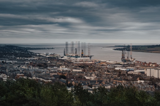 A coastal cityscape featuring a dense arrangement of buildings along the shoreline, with several large offshore drilling platforms visible in the water. The sky is overcast, casting a moody atmosphere over the landscape.