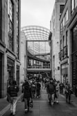 A bustling urban shopping area with people walking along a paved walkway between modern buildings. The architecture features a tall glass roof structure, creating an open, airy atmosphere. The facade of several shops, including a visible store named 'Next', lines the pathway.