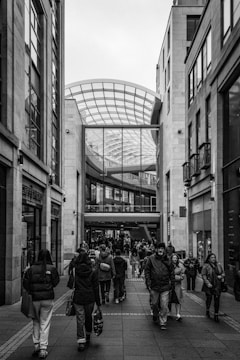 A bustling urban shopping area with people walking along a paved walkway between modern buildings. The architecture features a tall glass roof structure, creating an open, airy atmosphere. The facade of several shops, including a visible store named 'Next', lines the pathway.