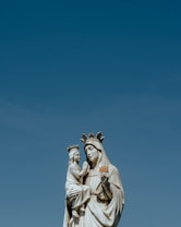 A marble statue depicts a crowned woman holding a child, both figures draped in flowing garments. The statue stands against a clear blue sky, showcasing intricate details on the crowns and clothing.