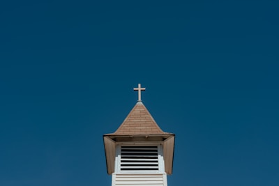 A church steeple with a cross on top against a clear blue sky. The steeple is made of wood with a shingled roof and features a series of horizontal vents just below the roof.