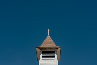 A church steeple with a cross on top against a clear blue sky. The steeple is made of wood with a shingled roof and features a series of horizontal vents just below the roof.