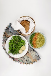 An arrangement of food items including a bowl of green soup garnished with herbs and fried onions, a plate of fresh green herbs with slices of lime, and a basket containing sliced brown bread, all placed on a floral-patterned cloth.