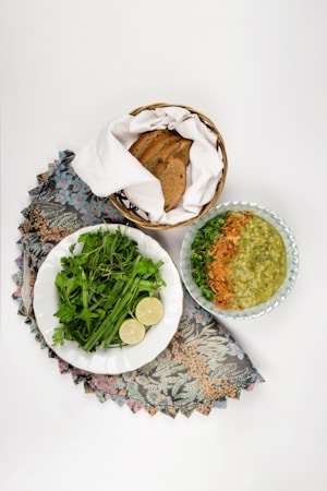 An arrangement of food items including a bowl of green soup garnished with herbs and fried onions, a plate of fresh green herbs with slices of lime, and a basket containing sliced brown bread, all placed on a floral-patterned cloth.