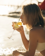 A person with blonde hair enjoys a cocktail by the beach, holding a glass with lemon and lime slices. The sunlight reflects off the water, creating a warm, serene setting.