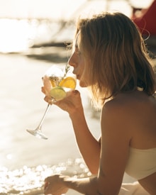A person with blonde hair enjoys a cocktail by the beach, holding a glass with lemon and lime slices. The sunlight reflects off the water, creating a warm, serene setting.
