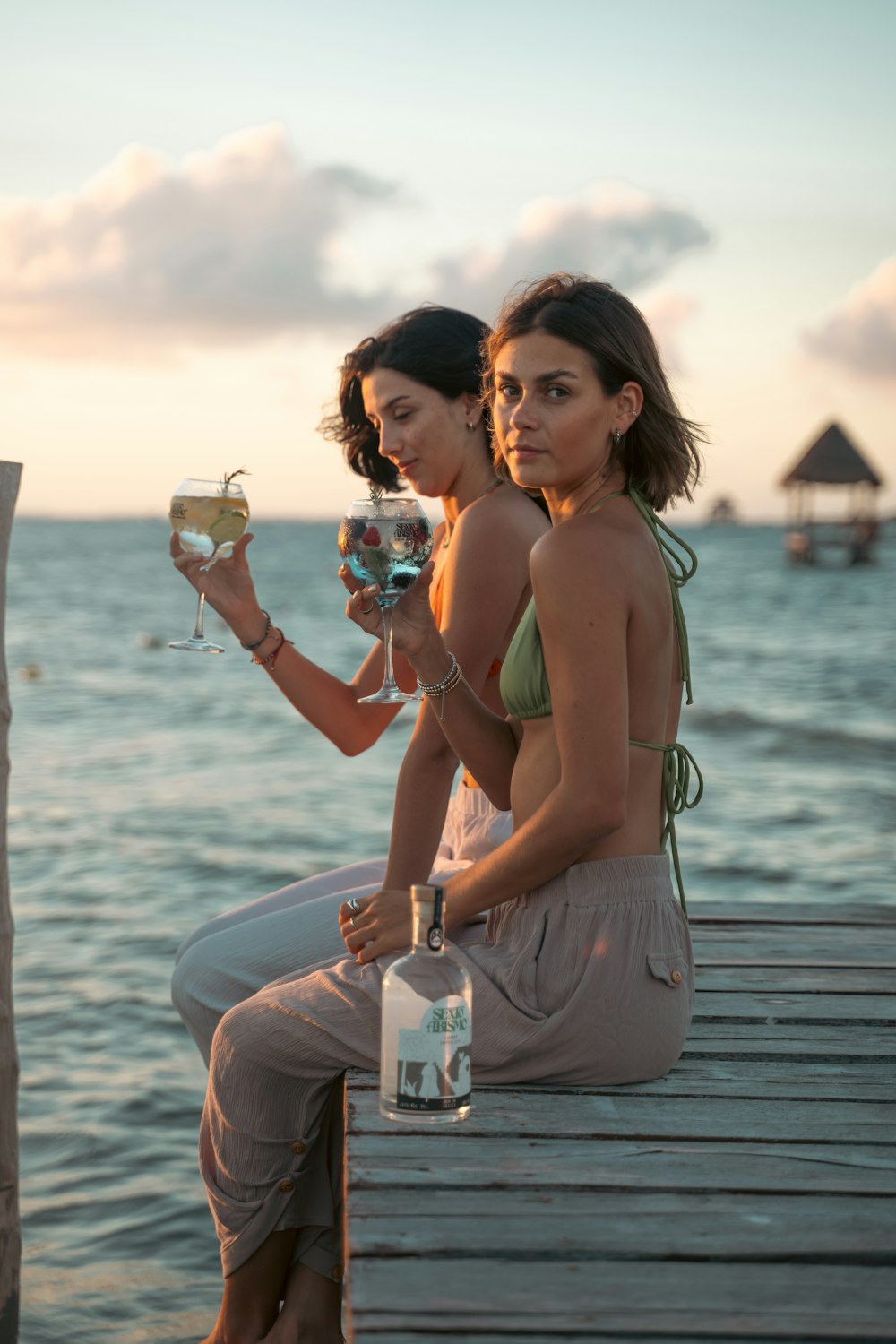 two women sitting on a dock holding wine glasses
